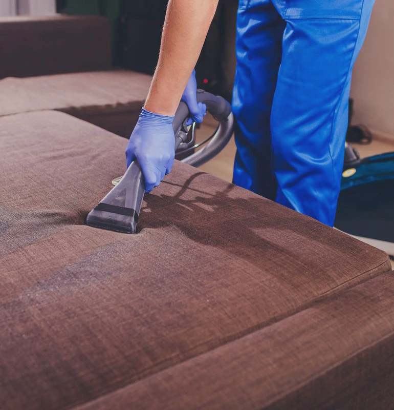 Pracownik w niebieskim stroju ochronnym i rękawiczkach czyści brązową kanapę